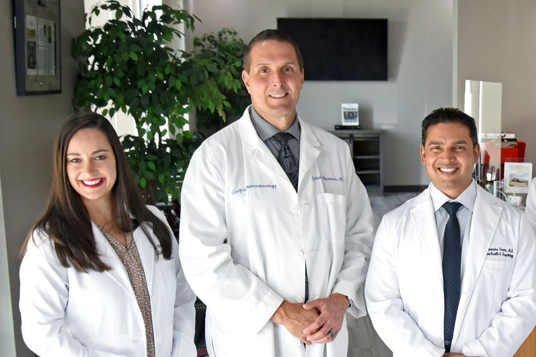 Three smiling medical professionals in white coats, likely a team of gastroenterologists, posing together in a clinical setting.