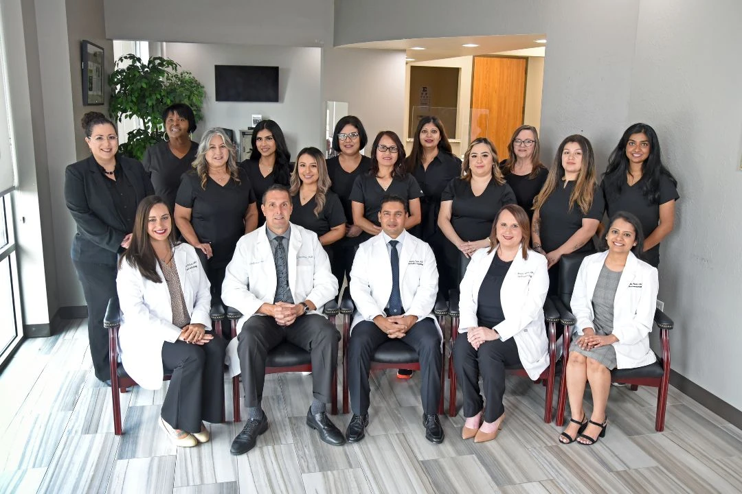 A group of medical professionals and staff, in white coats and black attire, pose for a team photo in a modern office setting.