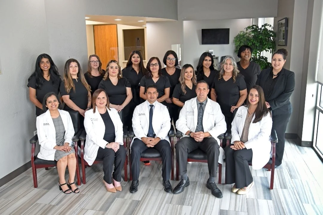 A group of medical professionals and staff, in white coats and black attire, pose for a team photo in a modern office setting.