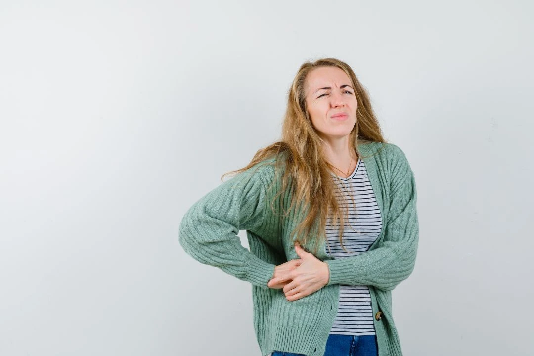 <br /> A woman grimacing in discomfort, holding her stomach, possibly experiencing abdominal pain or gastrointestinal distress.